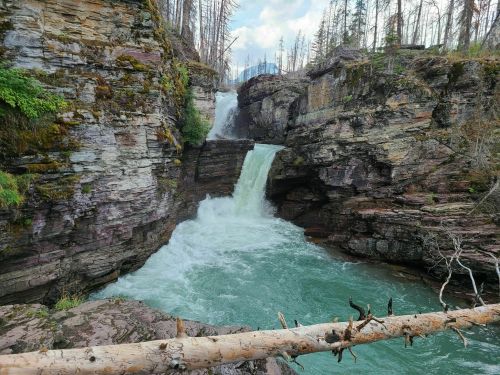 A scenic view of a waterfall cascading between rocky cliffs, surrounded by trees and a turquoise river.