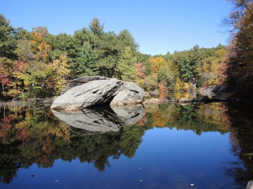 A serene lake scene with a large rock and colorful autumn trees reflecting in the calm water under a clear blue sky.