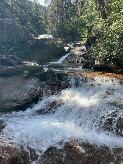 A serene waterfall cascades over rocky terrain, surrounded by lush green trees and a clear blue sky.