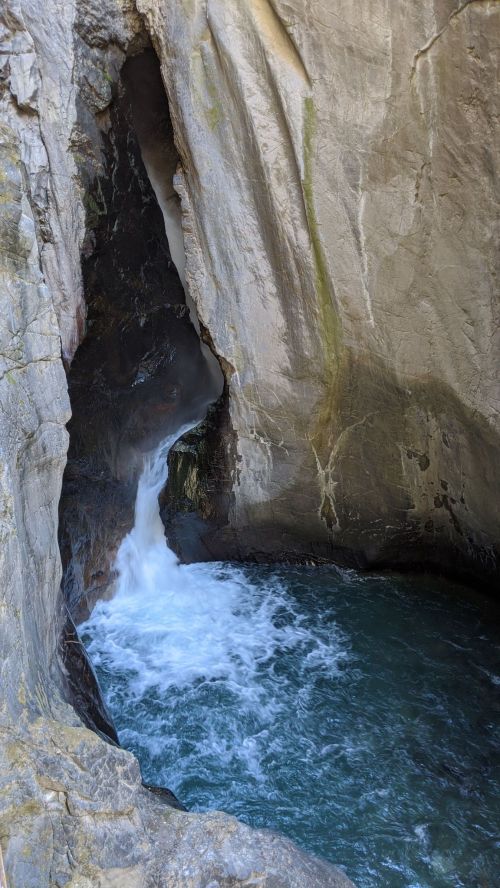 A small waterfall cascades into a rocky crevice, creating a pool of water below surrounded by steep stone walls.