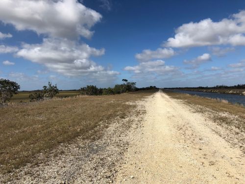 A dirt path stretches alongside a calm waterway, with grassy fields and scattered trees under a blue sky with clouds.
