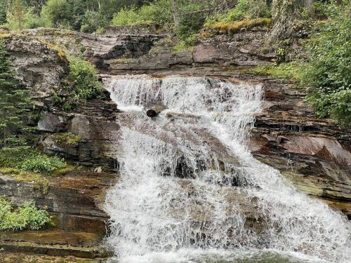 A cascading waterfall flows over rocky terrain, surrounded by lush greenery and trees.