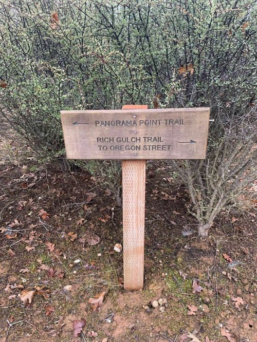 Wooden signpost indicating directions to Panorama Point Trail and Rich Gulch Trail to Oregon Street, surrounded by shrubs.