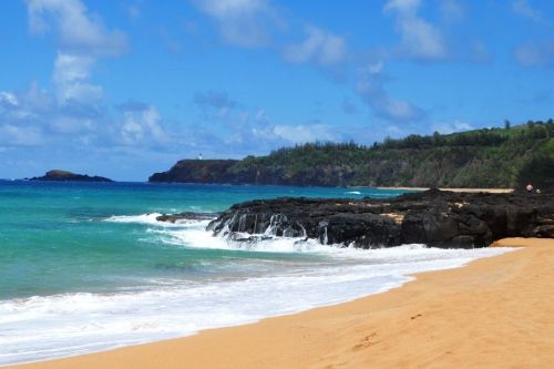 A serene beach scene with golden sand, gentle waves, and lush green hills under a blue sky with fluffy clouds.