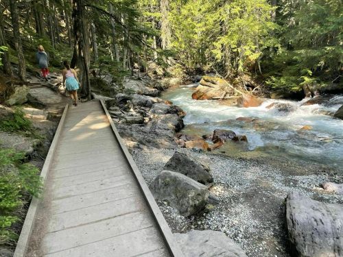 A wooden path alongside a flowing river, surrounded by lush greenery and rocky terrain, with two people walking.