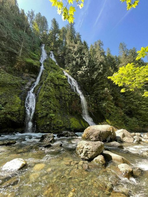 A serene waterfall cascades down a mossy cliff, surrounded by lush greenery and rocky riverbanks under a clear blue sky.