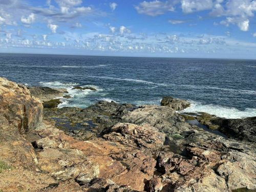 A rocky coastline with waves crashing against the shore under a blue sky with scattered clouds.