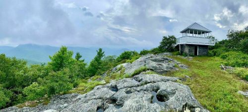 A scenic view of a rocky landscape with a lookout tower surrounded by greenery and distant mountains under a cloudy sky.