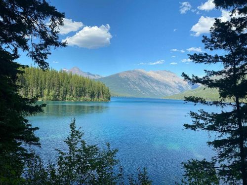 A serene lake surrounded by lush trees and mountains under a clear blue sky with fluffy clouds.