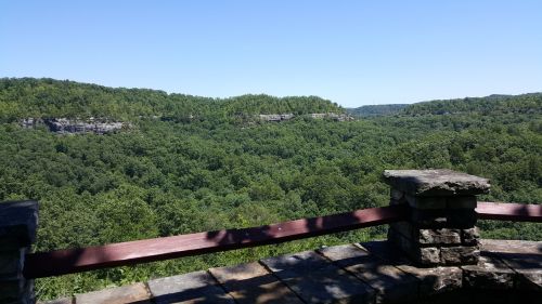 A scenic view of lush green hills and valleys under a clear blue sky, framed by a stone railing.