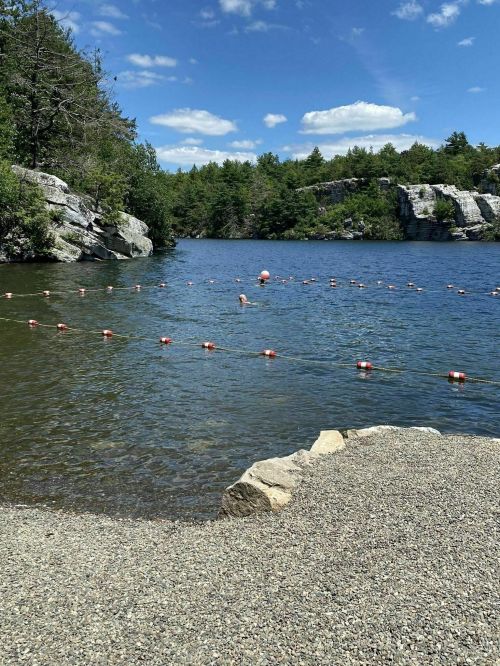 A serene lake surrounded by trees and rocky cliffs, with a sandy shore and floating buoys in the water.