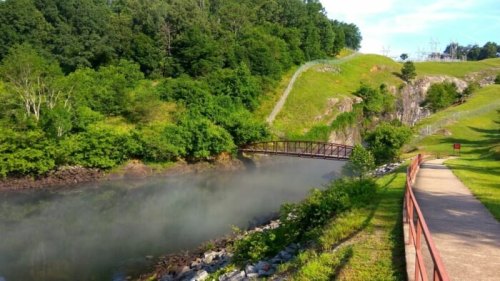 A serene landscape featuring a river, a small bridge, and lush greenery under a clear blue sky.