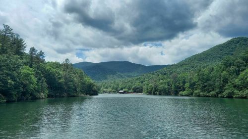 A serene lake surrounded by lush green trees and mountains under a cloudy sky.