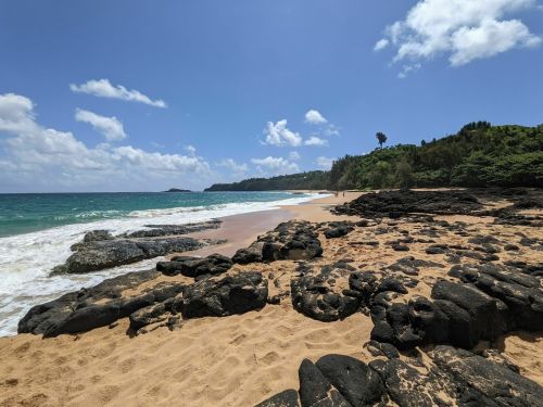 A scenic beach with rocky formations, golden sand, and gentle waves under a bright blue sky with scattered clouds.