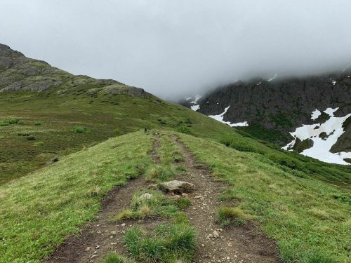A winding dirt path leads through green hills under a cloudy sky, with patches of snow on the mountains in the background.