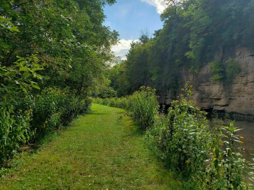 A serene path lined with greenery beside a calm river, flanked by rocky cliffs under a blue sky.