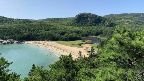 A scenic view of a sandy beach surrounded by lush green hills and a calm blue water inlet.