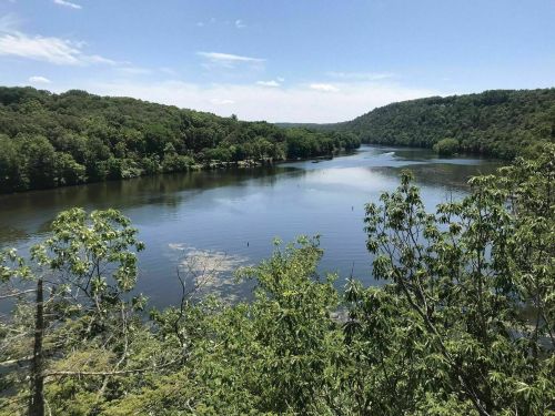 A serene river flows through lush green hills under a clear blue sky, reflecting the surrounding nature.