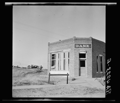 Abandoned brick bank building with broken windows, set against a barren landscape and machinery in the background.