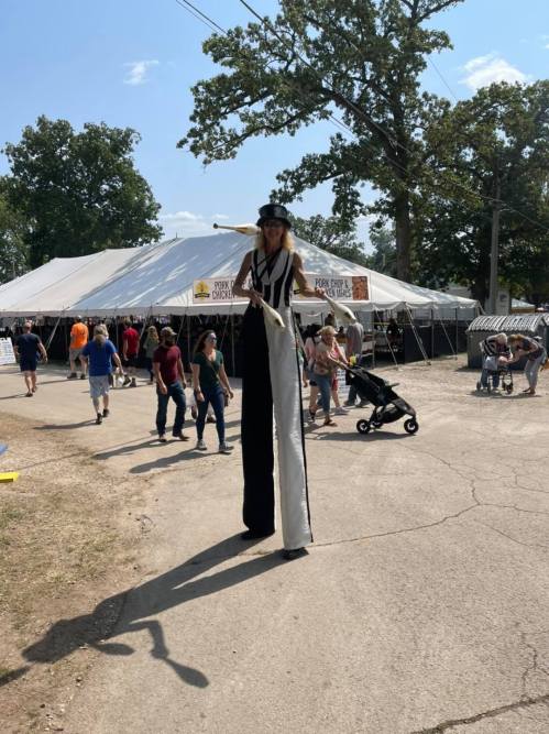 A tall stilt walker in a striped outfit entertains a crowd at an outdoor festival with tents in the background.