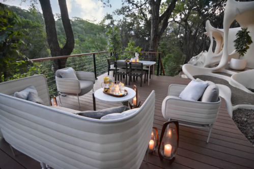 Cozy outdoor seating area with white furniture, a table, and lanterns, surrounded by lush greenery.