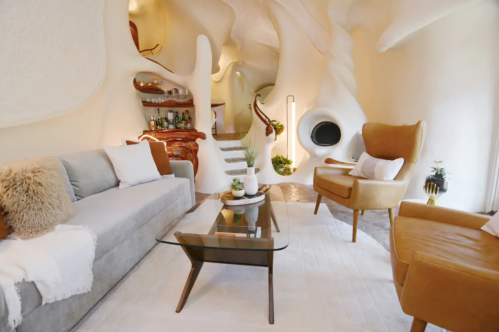 A modern, organic-shaped living room with a gray sofa, brown armchair, glass coffee table, and unique architectural details.