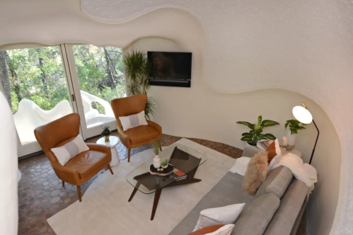 Cozy living room with curved walls, two brown chairs, a gray sofa, and a view of greenery through large windows.