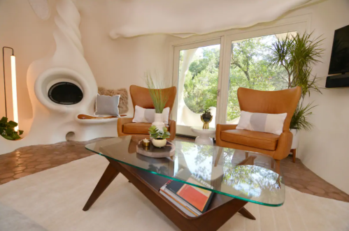 A modern living room with two brown chairs, a glass coffee table, and large windows overlooking greenery.