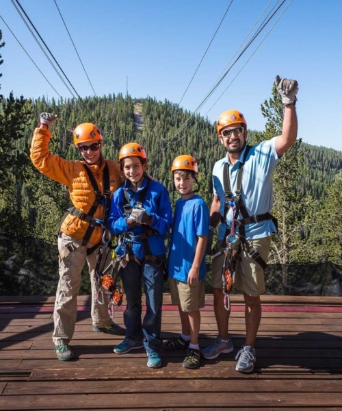 A family poses with smiles, wearing helmets and harnesses, ready for a zipline adventure in a forested area.