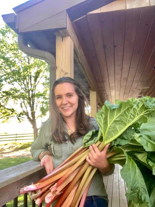 A woman smiles while holding a large bunch of rhubarb on a porch surrounded by greenery.