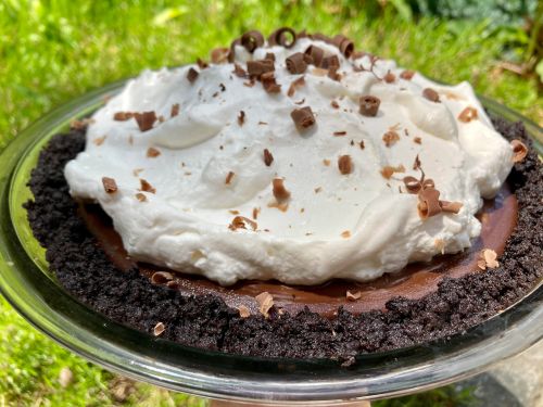 A chocolate pie with a whipped cream topping and chocolate shavings, set on a glass dish against a green background.