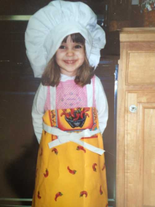 A smiling young girl in a chef's hat and colorful apron, standing in a kitchen.