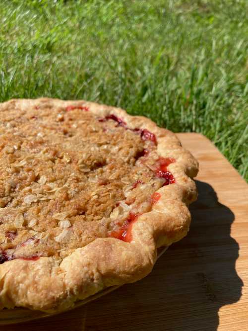 A freshly baked pie with a golden crust and crumb topping, resting on a wooden surface with green grass in the background.