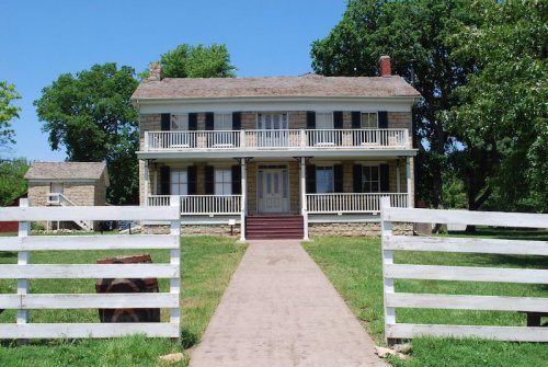 A two-story historic house with a front porch, surrounded by green grass and trees, with a white fence in the foreground.