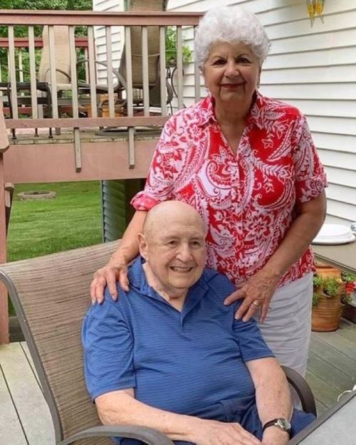 An elderly man sitting in a chair, smiling, with a woman standing behind him, both outdoors on a sunny day.