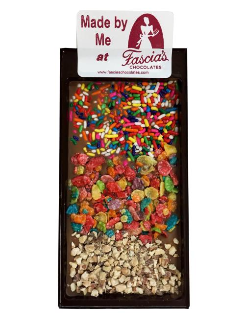 A chocolate bar topped with colorful sprinkles, fruity cereal, and crushed nuts, labeled "Made by Me at Fascía's Chocolates."