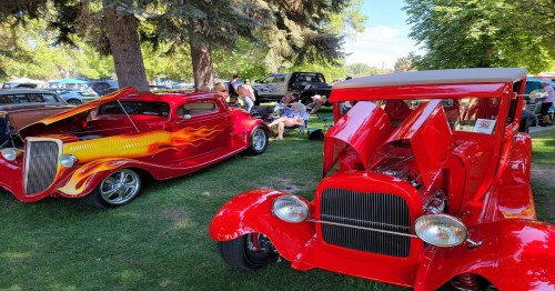 Two classic cars with vibrant red and flame designs parked in a grassy area, surrounded by trees and people enjoying the event.