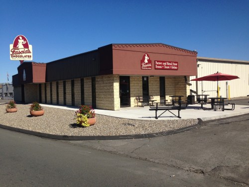Exterior view of a chocolate shop with a sign, outdoor seating, and flower pots, set in a sunny location.
