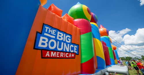 Colorful inflatable structures with "The Big Bounce America" logo under a blue sky with fluffy clouds.