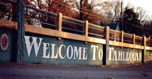 A sign reading "Welcome to Tahlequah" on a wall, surrounded by trees in a rural setting.
