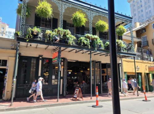A lively street scene featuring a historic building with greenery, people walking, and shops in a vibrant urban setting.