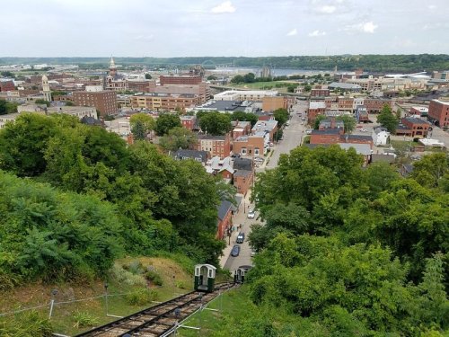 A scenic view of a city from a hilltop, featuring buildings, trees, and a railway track leading down.