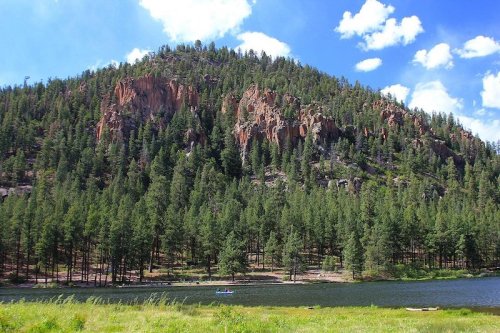 A serene landscape featuring a forested mountain, rocky cliffs, and a calm river under a blue sky with fluffy clouds.
