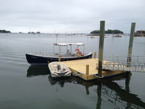 A small boat docked at a wooden pier on a calm, cloudy day with distant boats on the water.