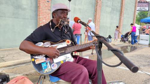 A musician plays guitar and sings at an outdoor event, surrounded by people and colorful stalls.