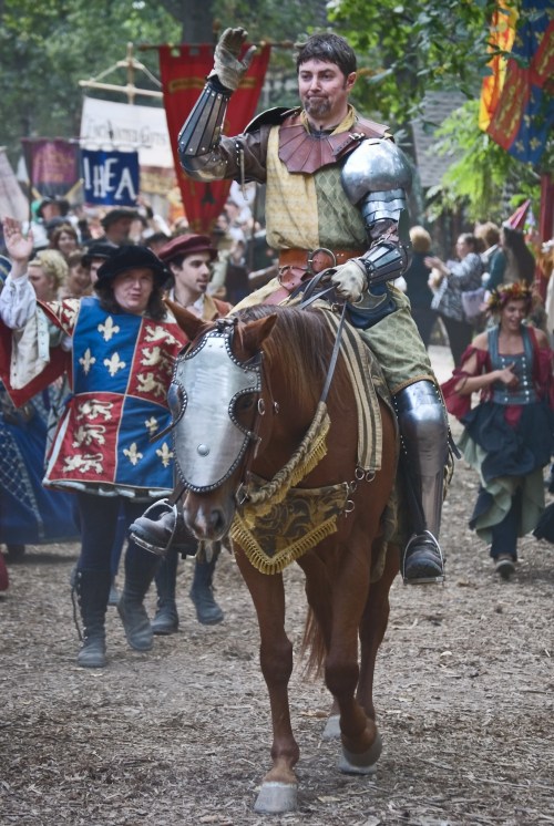 A knight in armor waves while riding a horse, surrounded by a festive crowd in medieval attire.