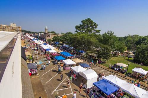 Aerial view of a vibrant outdoor market with colorful tents and people browsing among trees and buildings.