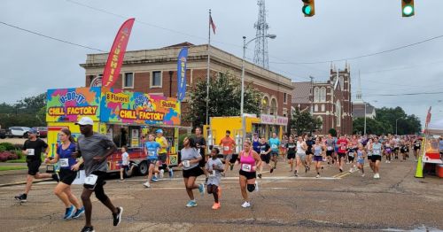 A large group of runners participates in a race, passing food vendors and a historic building on a cloudy day.