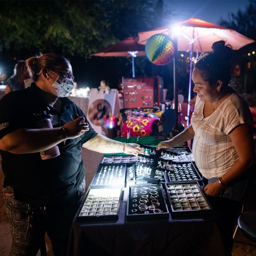 Two women interact at a night market, examining jewelry displayed on a table under soft lighting.