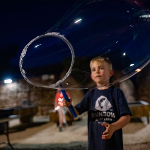 A young boy smiles as he holds a bubble wand, creating a large bubble against a nighttime backdrop.